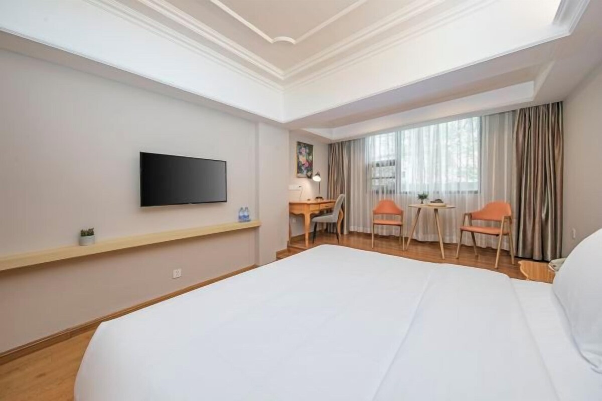 A hotel room with a large bed, a desk, two chairs, a TV on the wall, and a window with curtains.