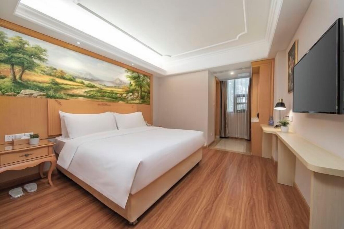 A hotel room with a bed, wooden flooring, a painting on the wall, a desk, and a TV.
