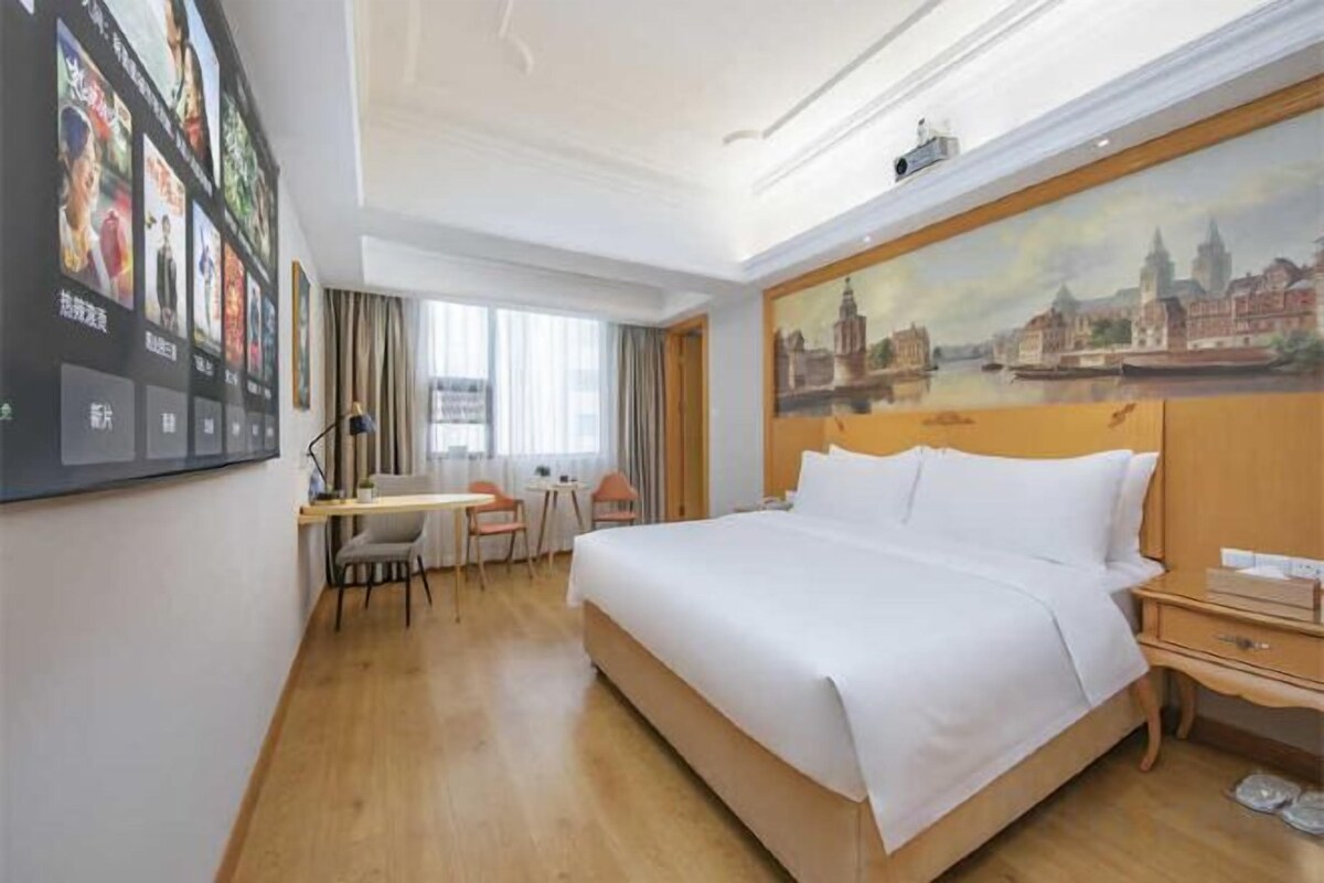A hotel room with a bed, desk, chair, and a painting on the wall.