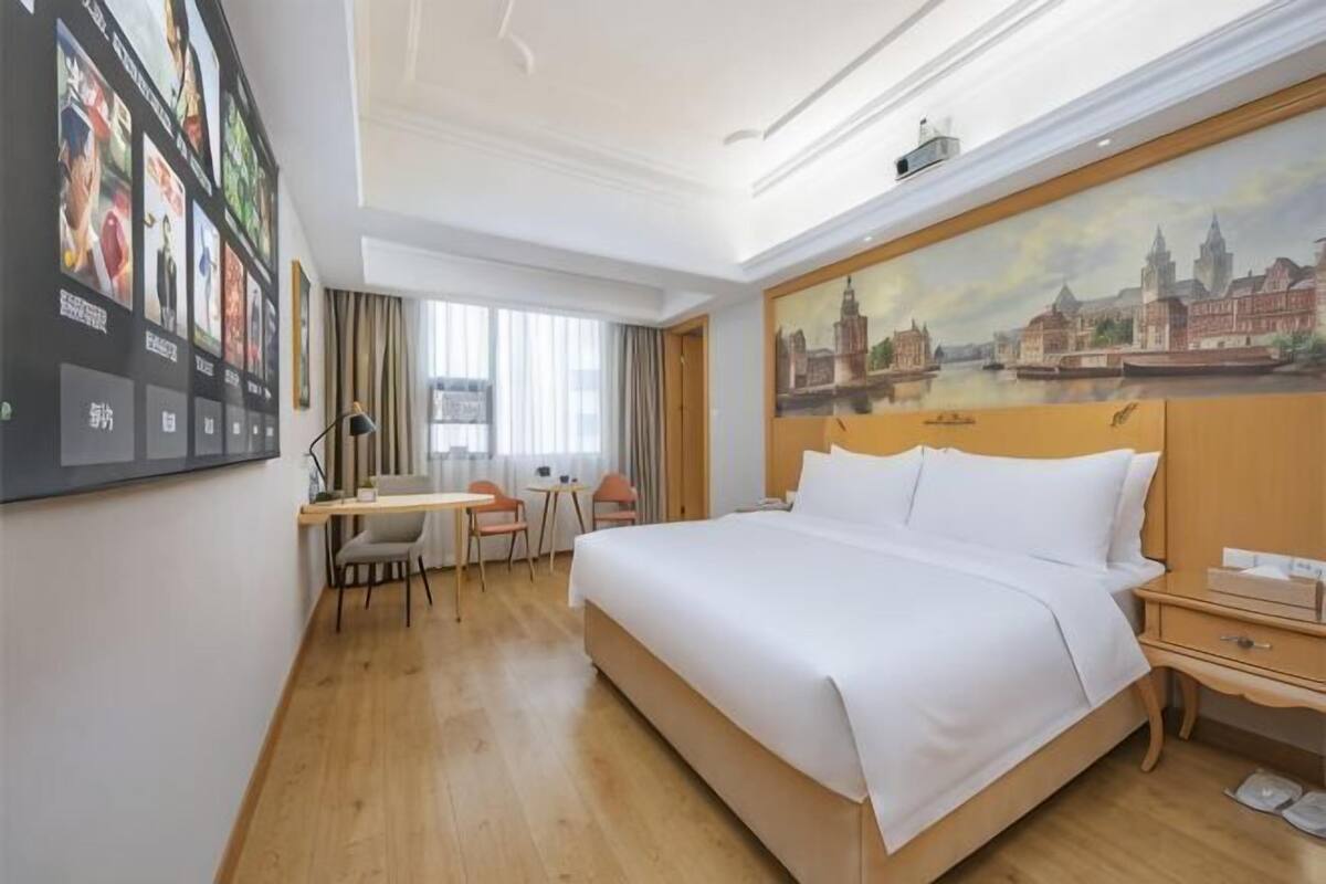A hotel room with a bed, desk, chair, and a painting of a cityscape.