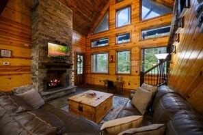 A log cabin interior with a stone fireplace, leather sofa, and large windows. - Whistling Duck · Stocked Pond: Gameroom, Shuffleboard, & Hot Tub (Broken Bow)