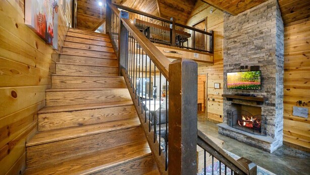 A wooden staircase leading to a second floor with a stone fireplace and a television on the wall. - Whistling Duck · Stocked Pond: Gameroom, Shuffleboard, & Hot Tub (Broken Bow)