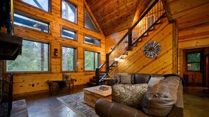 A log cabin interior with a staircase, leather sofa, wooden walls, and large windows. - Whistling Duck · Stocked Pond: Gameroom, Shuffleboard, & Hot Tub (Broken Bow)