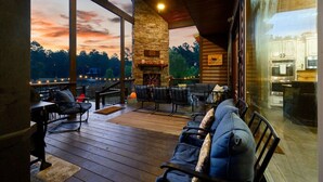 A spacious outdoor deck with wooden flooring, a stone fireplace, and a view of the surrounding landscape. - Whistling Duck · Stocked Pond: Gameroom, Shuffleboard, & Hot Tub (Broken Bow)