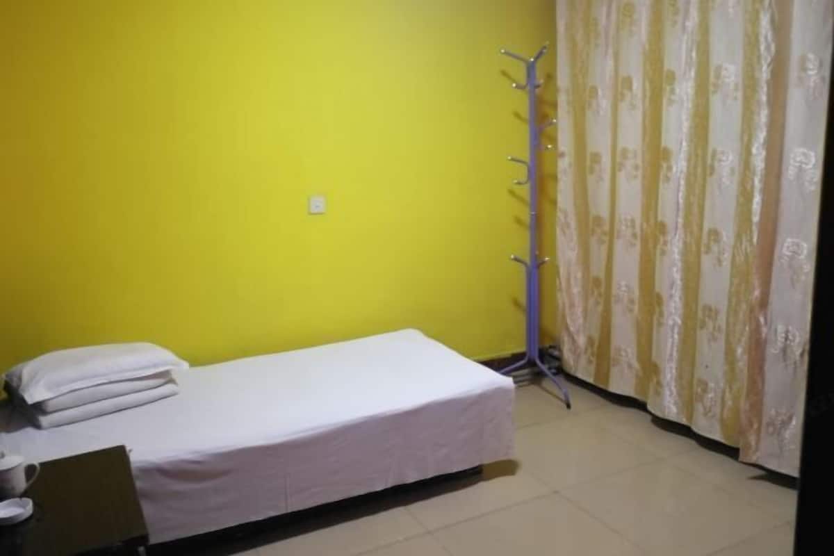 Ein kleines Zimmer mit einem Einzelbett, einer gelben Wand, einer weißen Bettdecke, einem Kleiderhaken und einem Vorhang mit Blumenmuster.