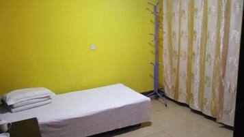 A small, single-bed room with a yellow wall, a white bedspread, a coat rack, and a curtain with a floral pattern.