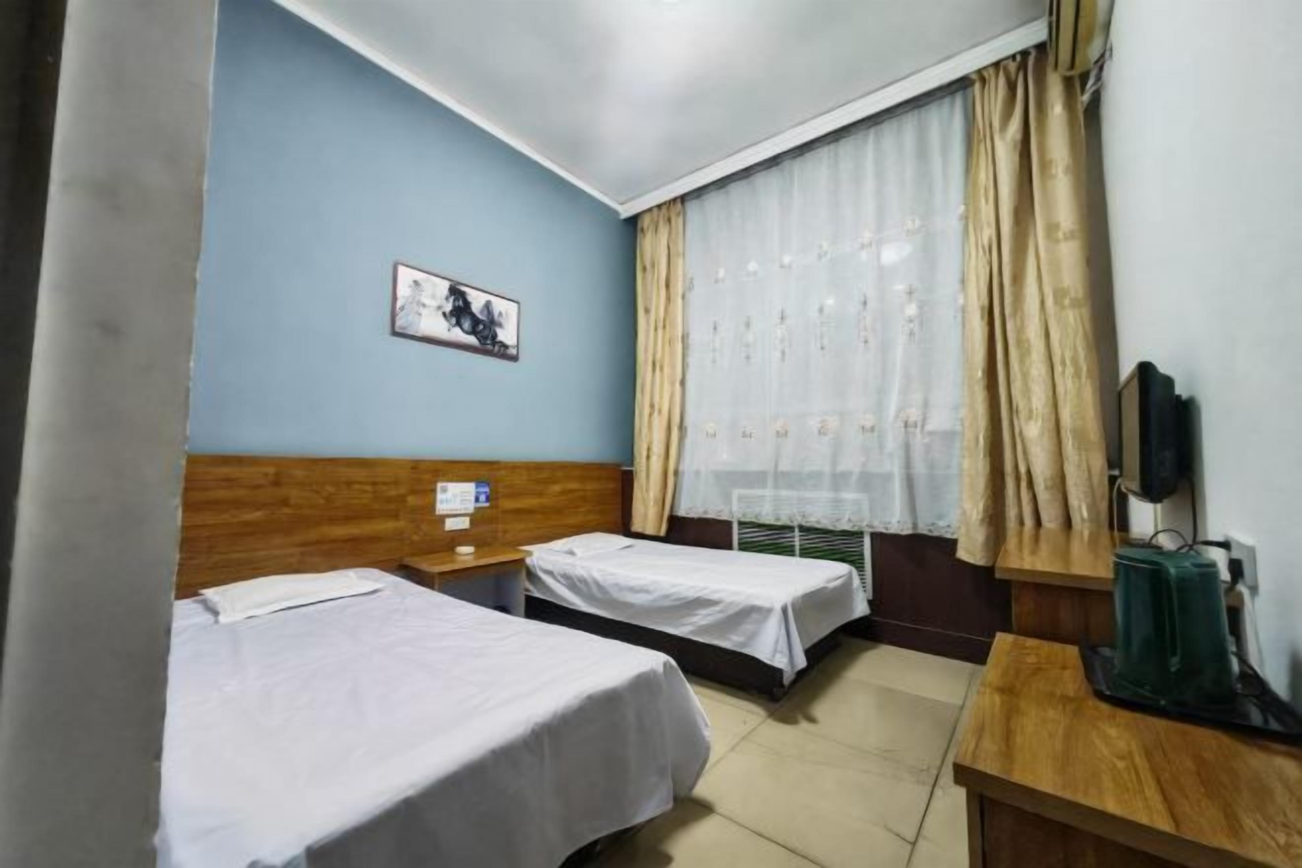 A hotel room with two beds, a wooden headboard, a small table, a TV, and a window with curtains.