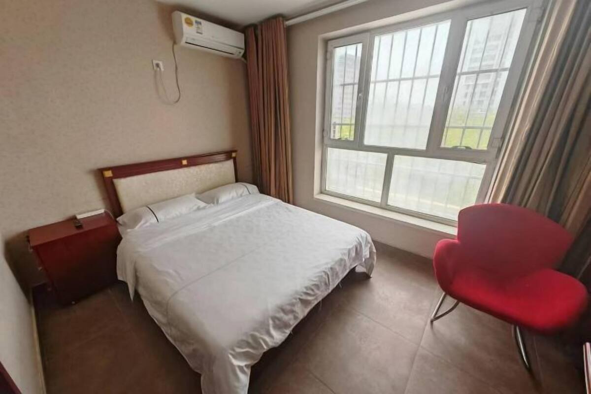 A hotel room with a bed, a red chair, a window with curtains, and an air conditioning unit.