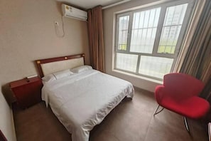 A hotel room with a bed, a red chair, a window with curtains, and an air conditioning unit.