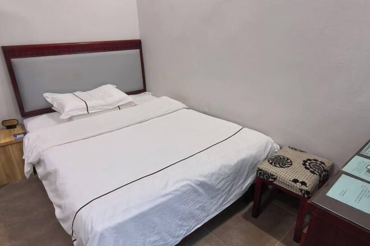A neatly made bed with a wooden headboard, a small bedside table with a lamp, and a patterned stool.