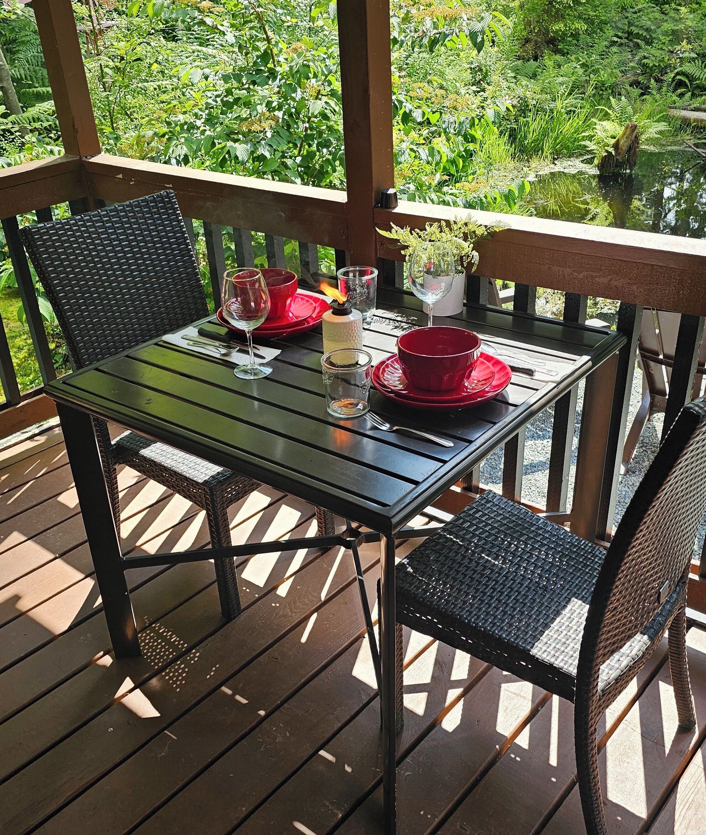 An outdoor wooden deck with a table set for two, featuring wicker chairs and red tableware.