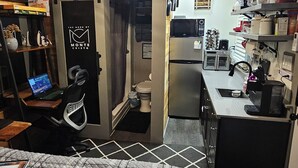 A compact room with a bed, a desk with a laptop, a kitchenette, and a ceiling fan.