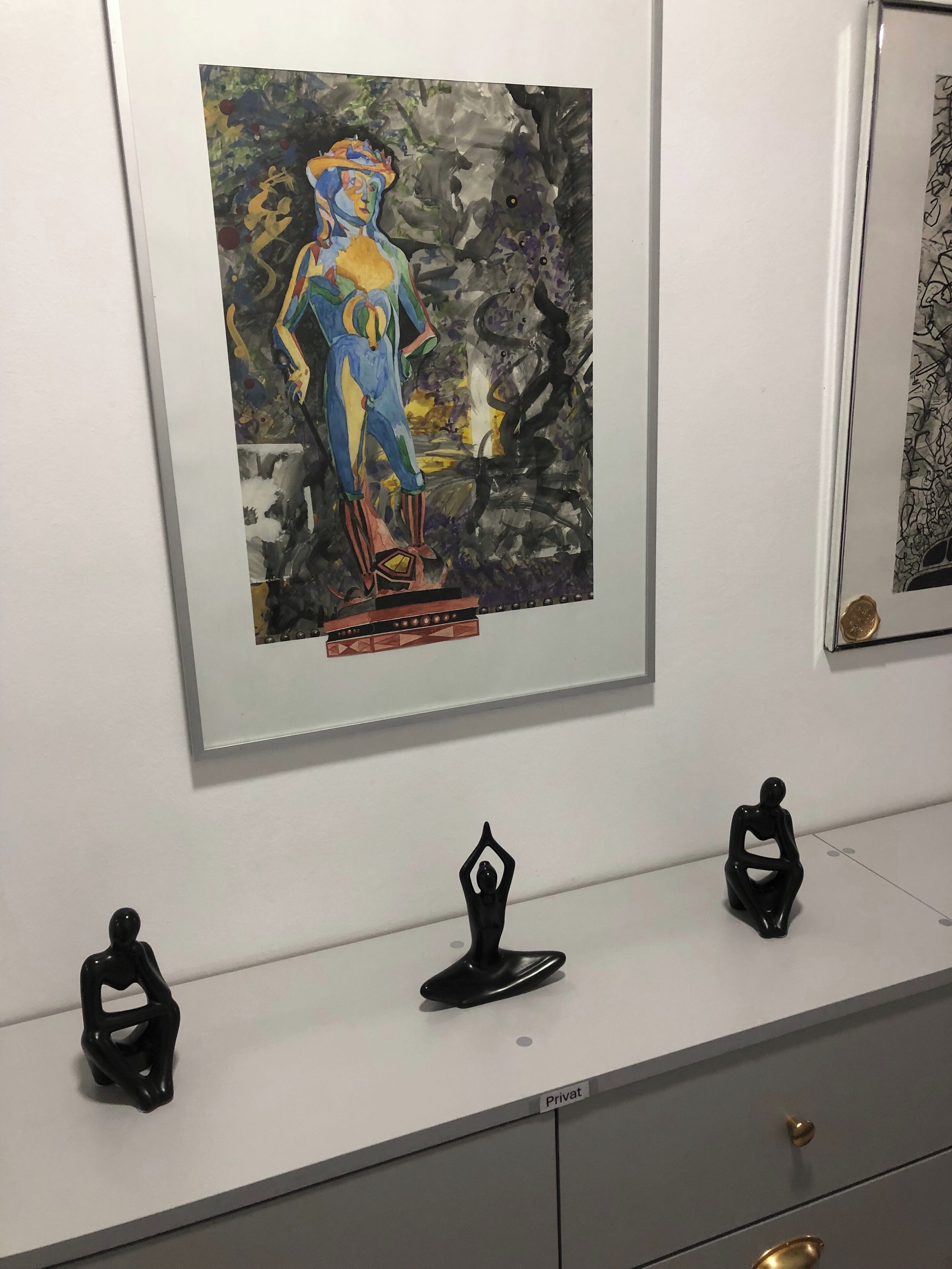 Une œuvre d’art encadrée accrochée au mur, une petite sculpture noire posée sur un comptoir et, en dessous, un meuble gris.