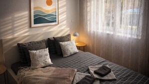 A bedroom with a bed, pillows, a bedside table with a lamp, a window with curtains, and a framed picture on the wall.