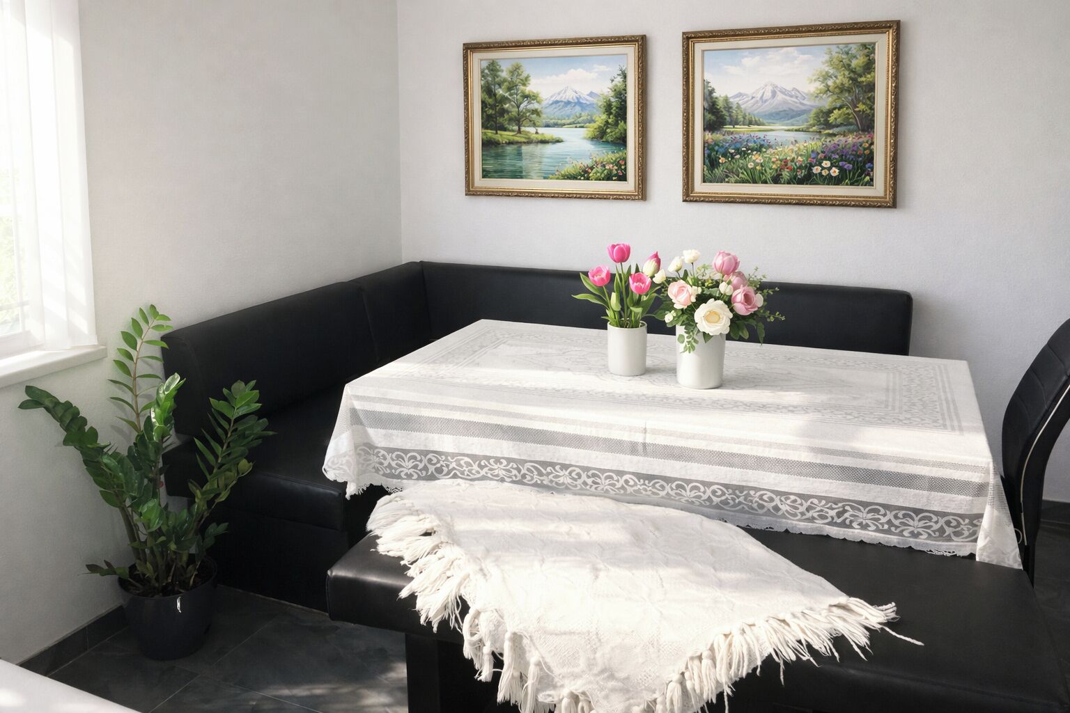 A room with a black leather bench, a white table with a lace table runner, two framed landscape paintings, and a potted plant.