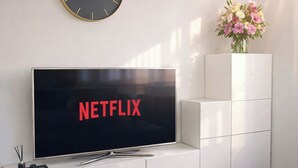 A modern living room with a flat-screen TV displaying the Netflix logo, a minimalist white entertainment unit, a wall clock, and a decorative vase with flowers.