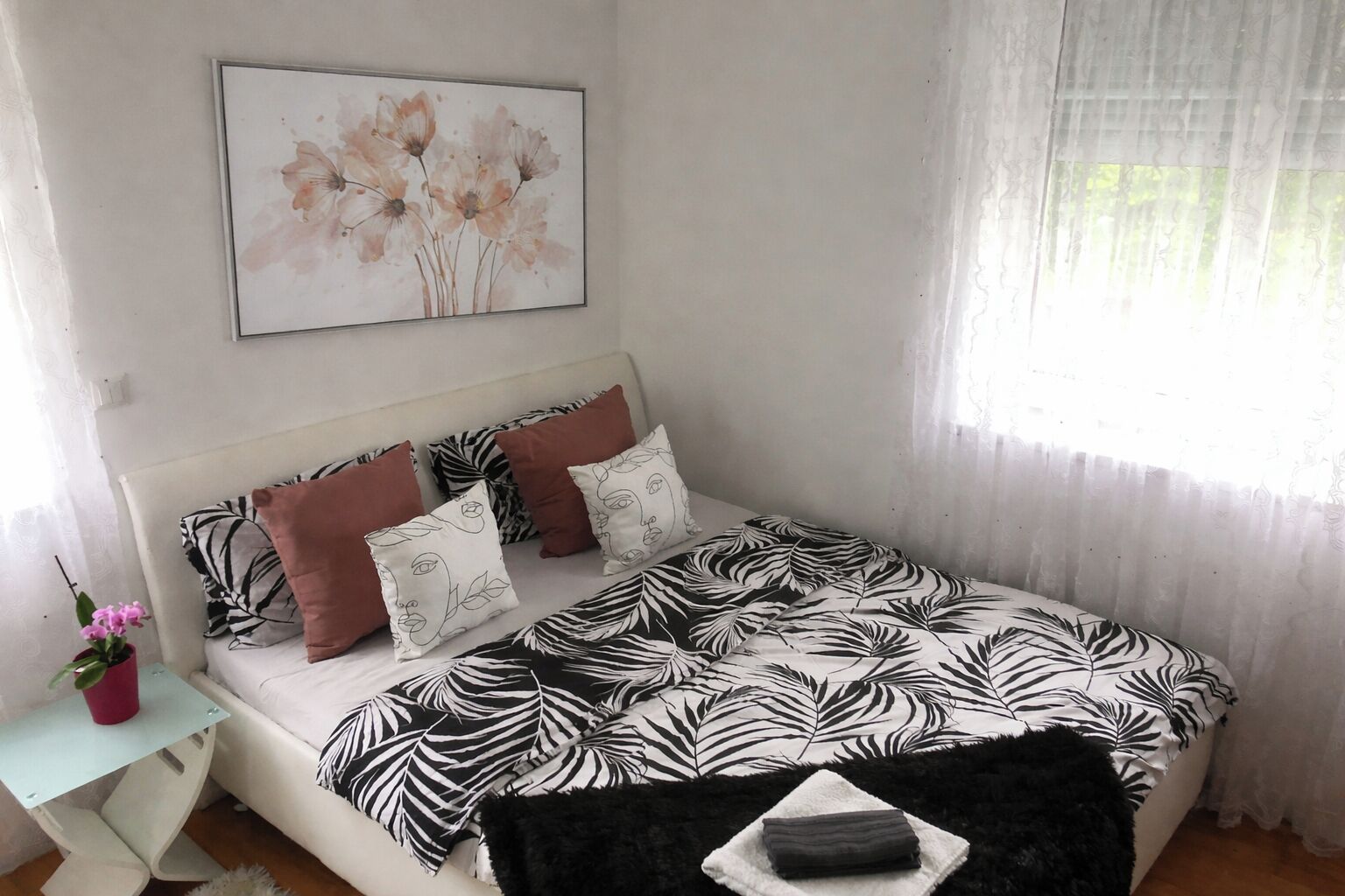 Un dormitorio con una cama, almohadas, una mesita de noche con una planta y una ventana con cortinas translúcidas.