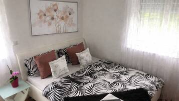 Un dormitorio con una cama, almohadas, una mesita de noche con una planta y una ventana con cortinas translúcidas.
