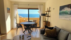 Un salon comprenant un canapé, une table basse et un coin repas avec des chaises et une table. De grandes fenêtres offrent une vue sur l’océan.
