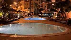 Un espace piscine aménagé pour la nuit, avec des chaises longues et des parasols, entouré d’immeubles dotés de balcons et de palmiers.