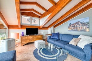 A modern living room with a blue sofa, wooden beams, a TV, and a large window.