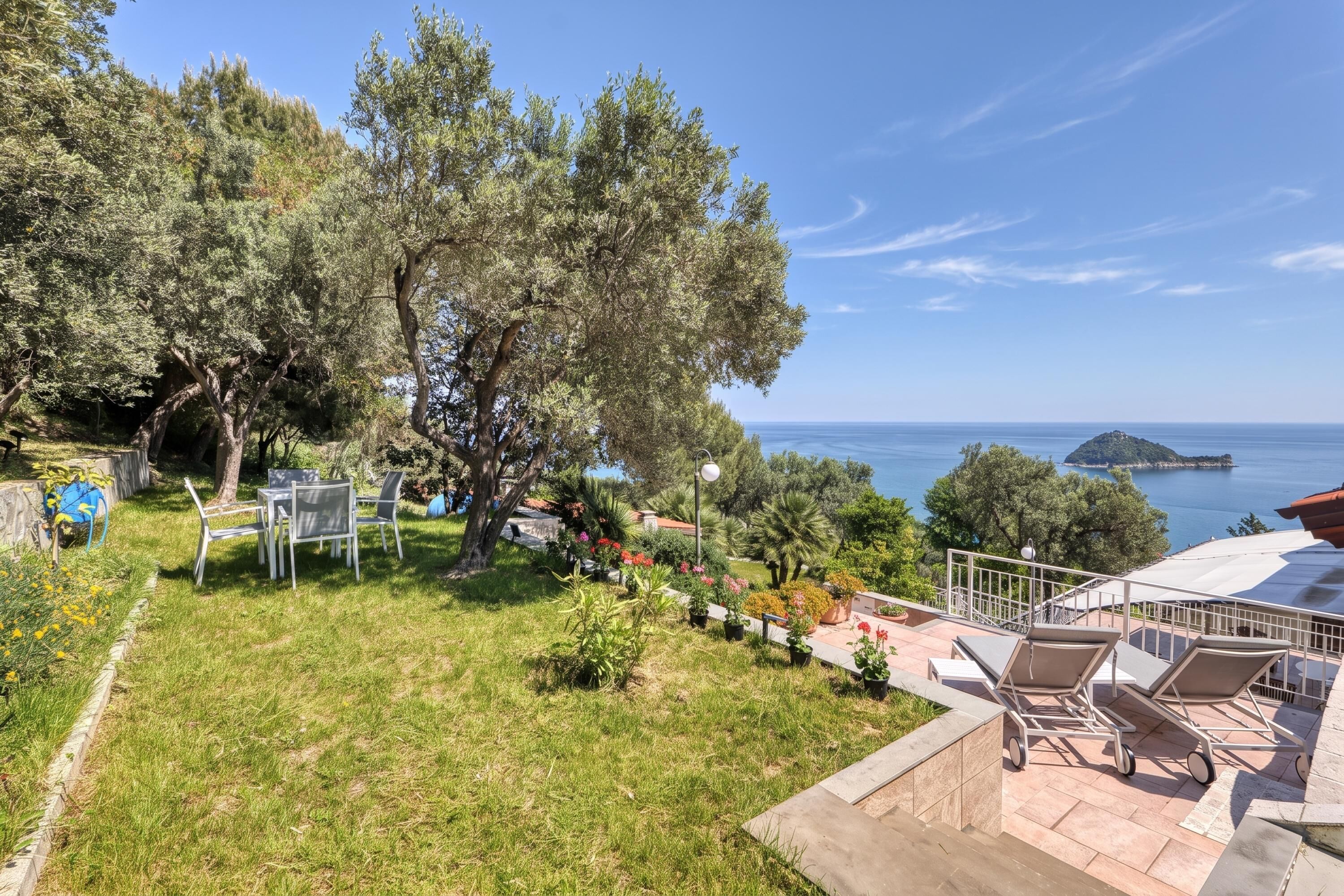 Una terrazza con mobili da esterno, un giardino con diverse piante e vista sul mare e un’isola in lontananza.
