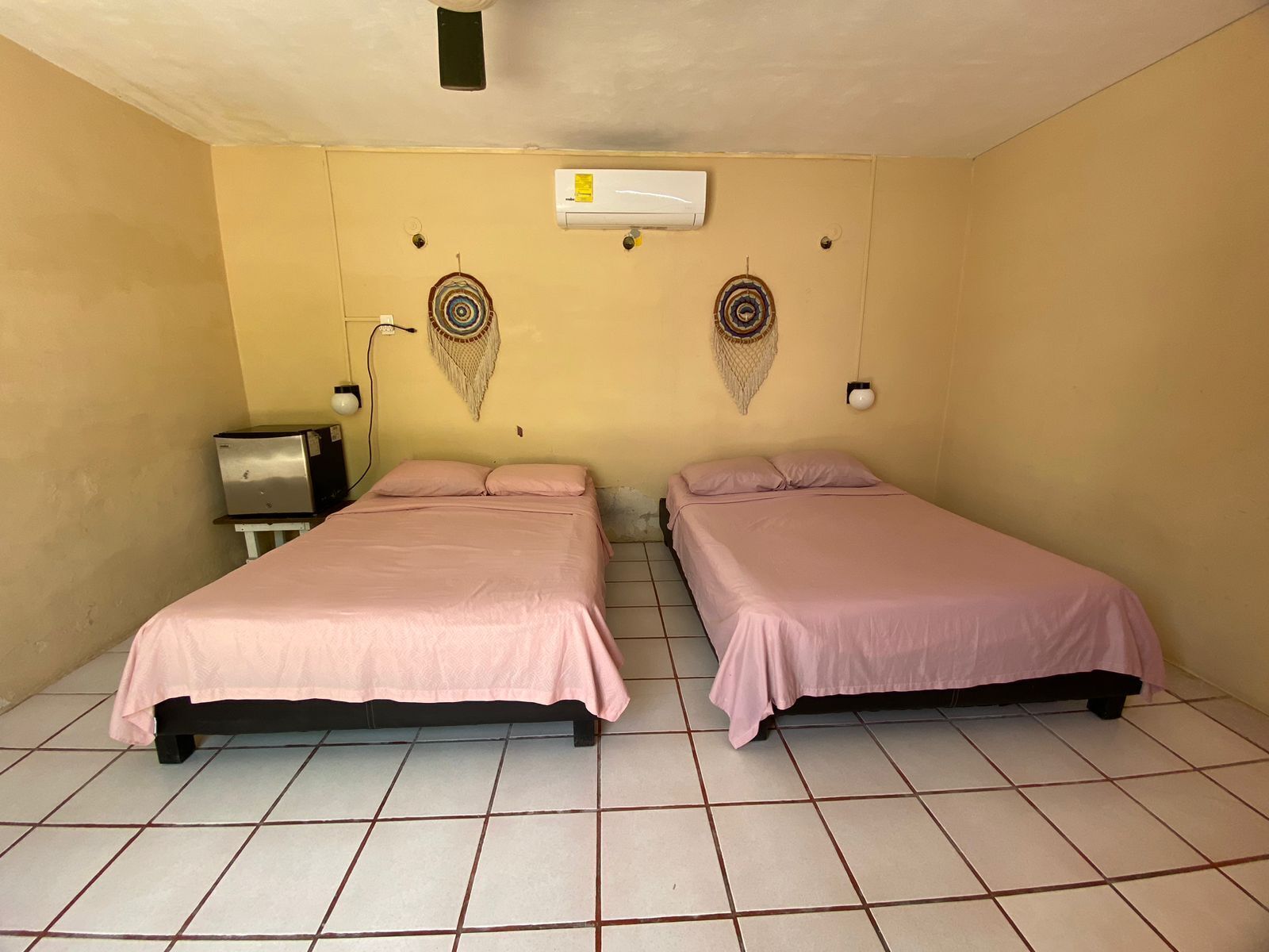 Una habitación pequeña y limpia con dos camas, aire acondicionado instalado en la pared y un escritorio pequeño con una lámpara.