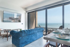 A modern living room with a blue sofa, a dining table set for two, and a large window overlooking the sea.