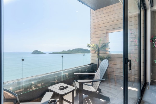 A balcony with a glass railing, a white chair, a small table, and a view of the sea and distant islands.