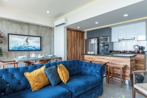 Una sala de estar moderna con un sofá azul, detalles de madera, una cocina con electrodomésticos de acero inoxidable y un comedor.
