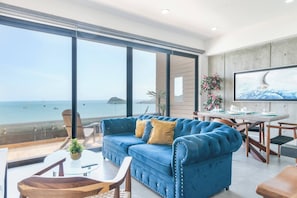 A modern living room with a blue sofa, a dining table, and a large window overlooking the sea.