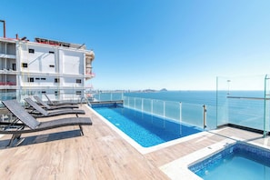 A rooftop pool with lounge chairs, a clear view of the sea, and a modern building in the background.