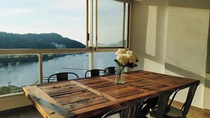 A wooden dining table with chairs and a vase of flowers, set against a large window overlooking a river and hills.