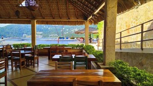 An outdoor dining area with wooden tables and chairs, a stone wall, and a view of a pool and mountains.