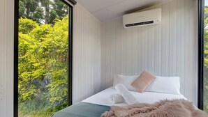 A modern bedroom with a large window, a bed with white linens, a fluffy beige throw, and an air conditioning unit on the wall. - Tiny Sublime by Tiny Away (Kia Ora)