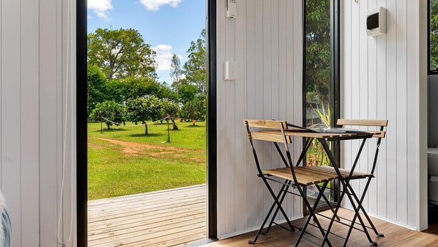 A room with a wooden table and chair, a view of a green landscape through a sliding door, and a white wall with a thermostat. - Tiny Sublime by Tiny Away (Kia Ora)