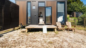 A modern, compact cabin with a wooden deck, two Adirondack chairs, and a sign indicating a 'Living Room'.