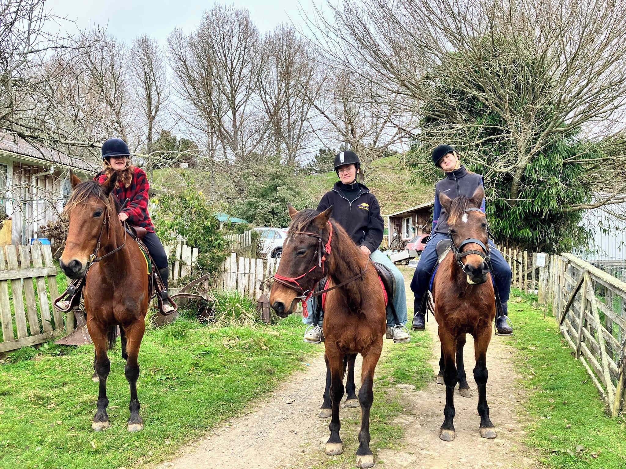 Trois personnes à cheval, sur un chemin de terre, avec une clôture en bois d’un côté et des arbres en arrière-plan.