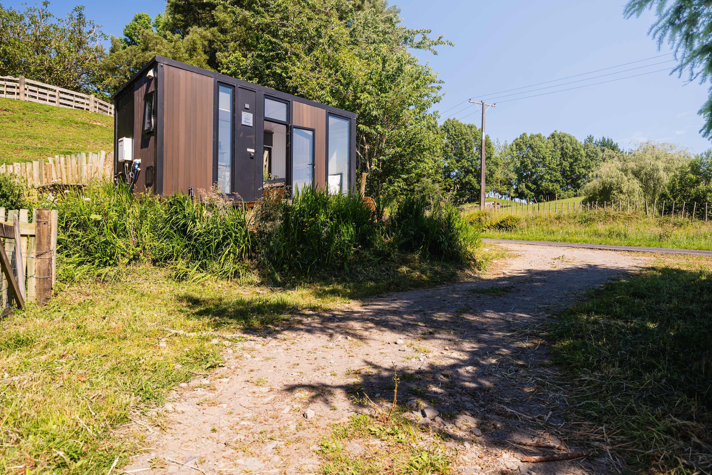 Une cabane moderne et compacte, dotée d’une grande porte coulissante, entourée d’une végétation luxuriante et accessible par un chemin de terre qui mène à une route.