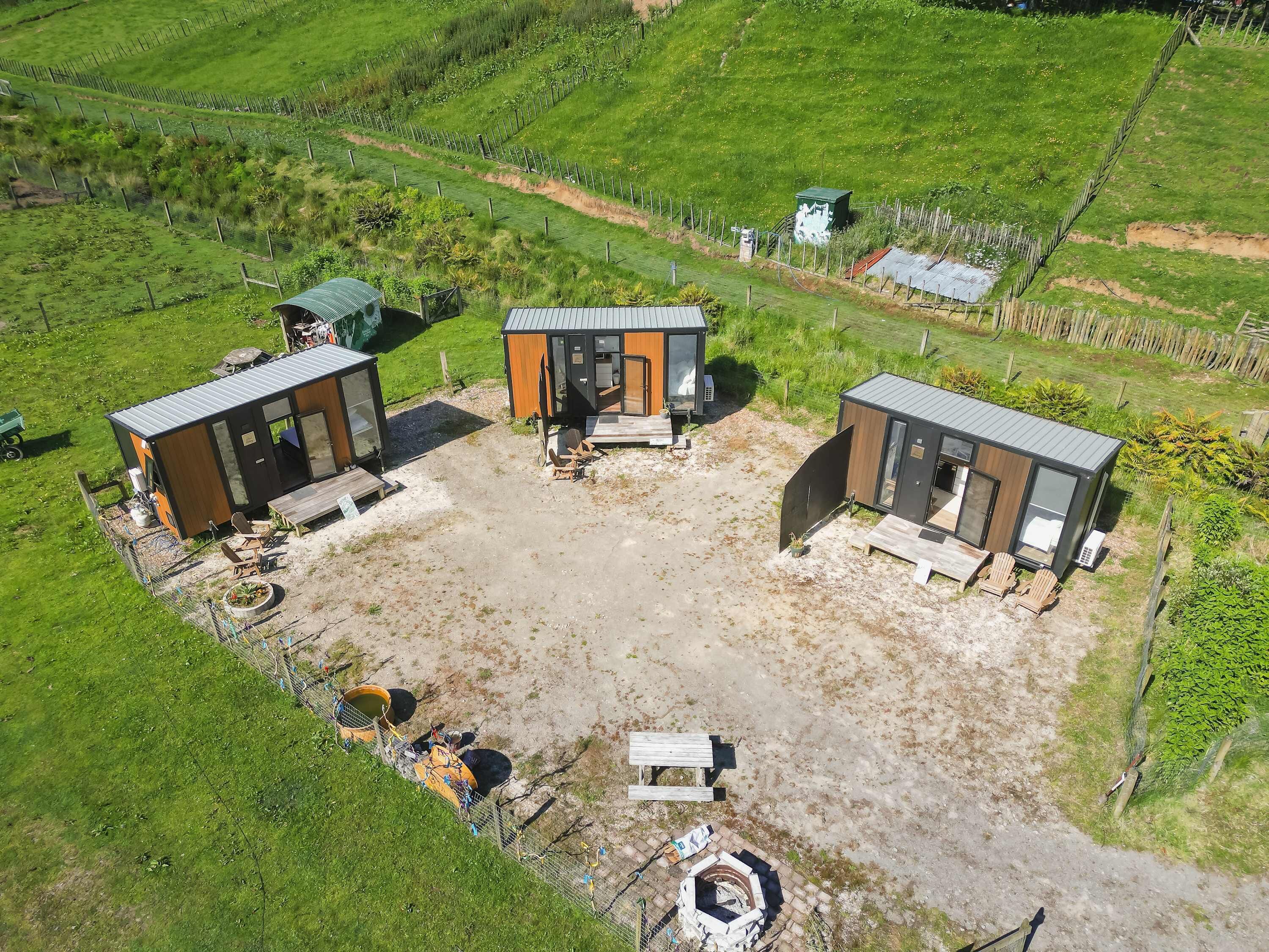 A modern, compact housing complex with three cabins, each featuring a large window and a door. The area includes a central open space with a fire pit and outdoor seating.
