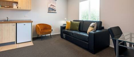 A compact living space with a kitchenette, a sofa with cushions, a glass table, and a chair.
