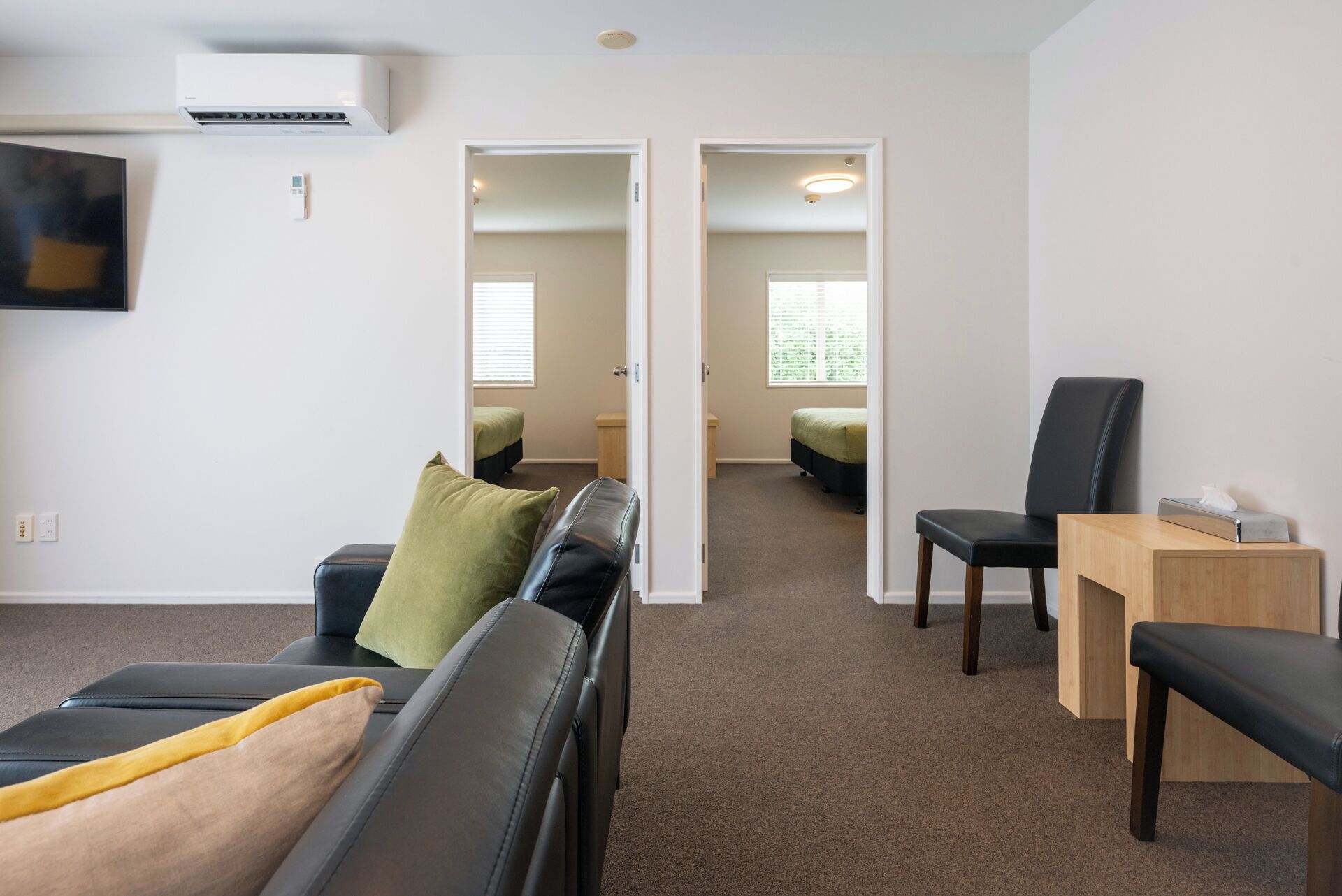 Une pièce moderne et bien éclairée, avec un canapé, un fauteuil, une petite table et un lit recouvert d’une couverture verte. Un téléviseur est fixé au mur et il y a une fenêtre avec des stores.