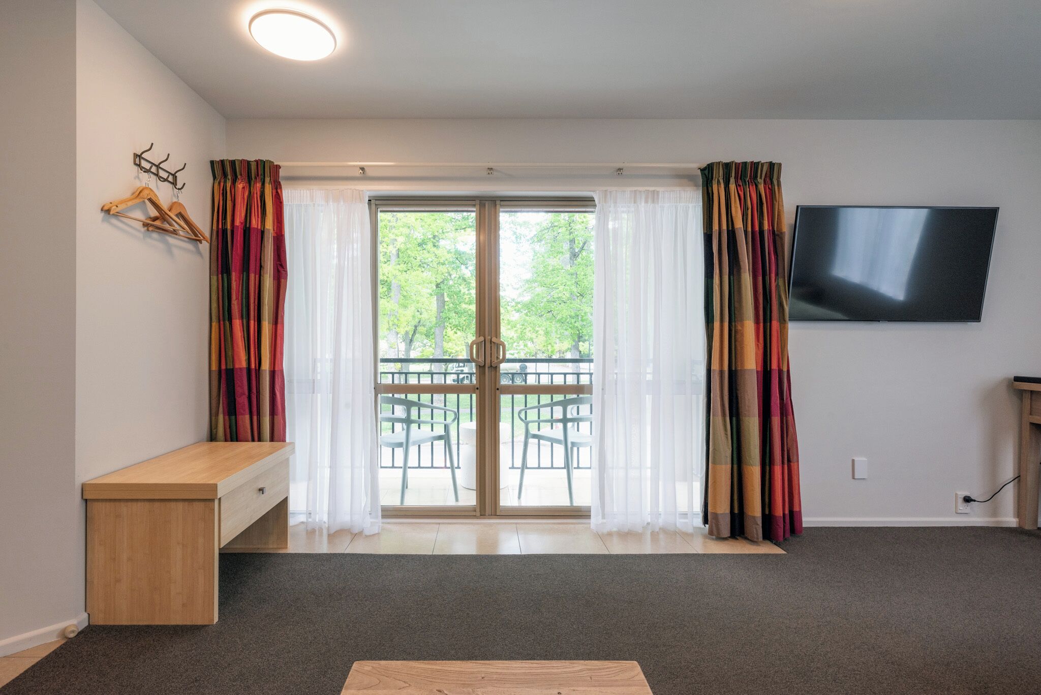 A room with a wooden bench, a TV mounted on the wall, and a sliding glass door leading to an outdoor area with chairs.