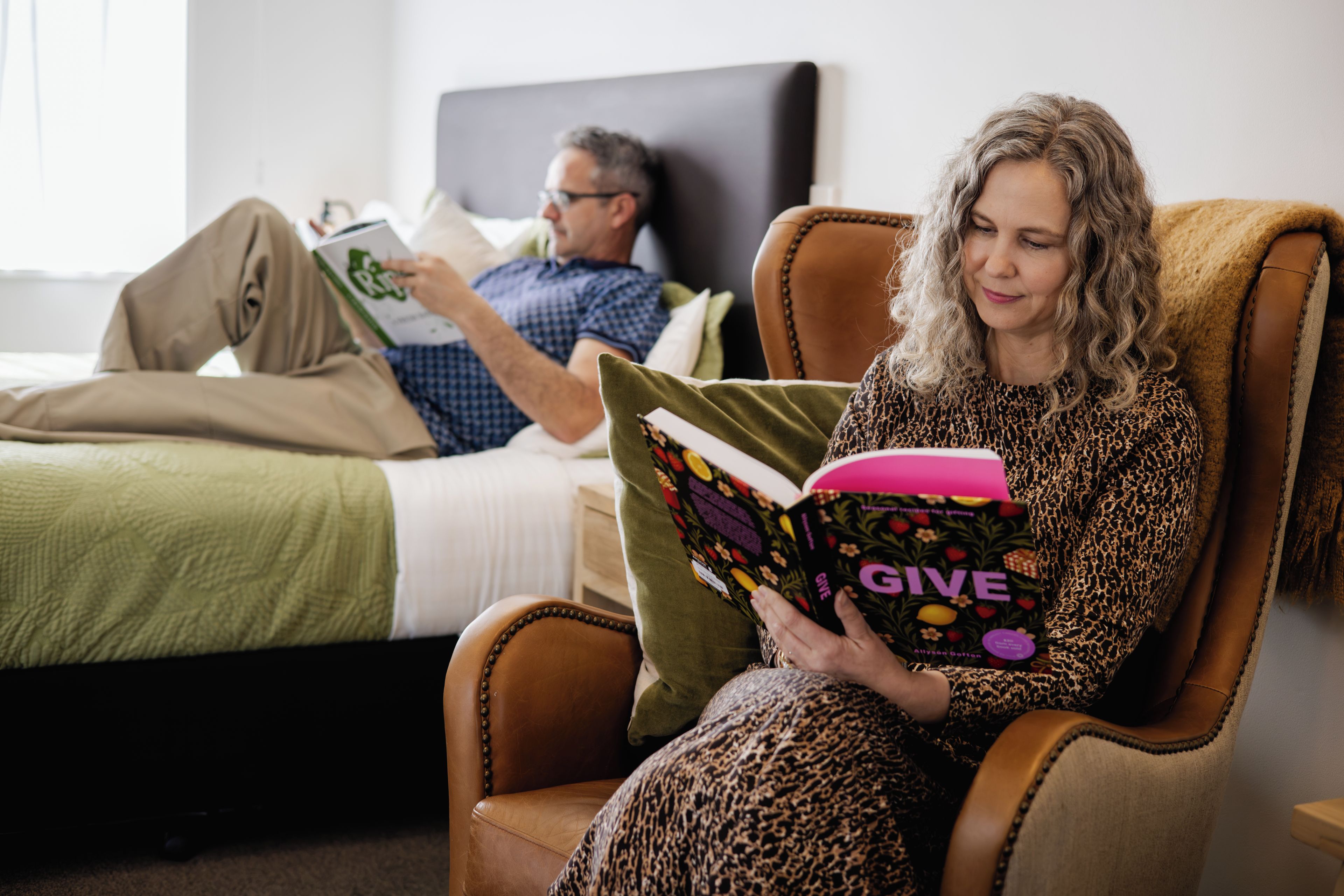 Eine Person sitzt in einem Sessel und liest ein Buch mit dem Titel 'GIVE' in einem Zimmer, das ein Bett und einen Nachttisch beinhaltet.