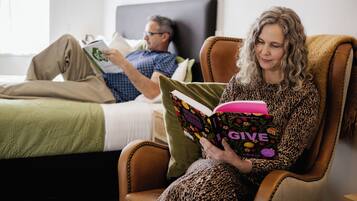 Eine Person sitzt in einem Sessel und liest ein Buch mit dem Titel 'GIVE' in einem Zimmer, das ein Bett und einen Nachttisch beinhaltet.