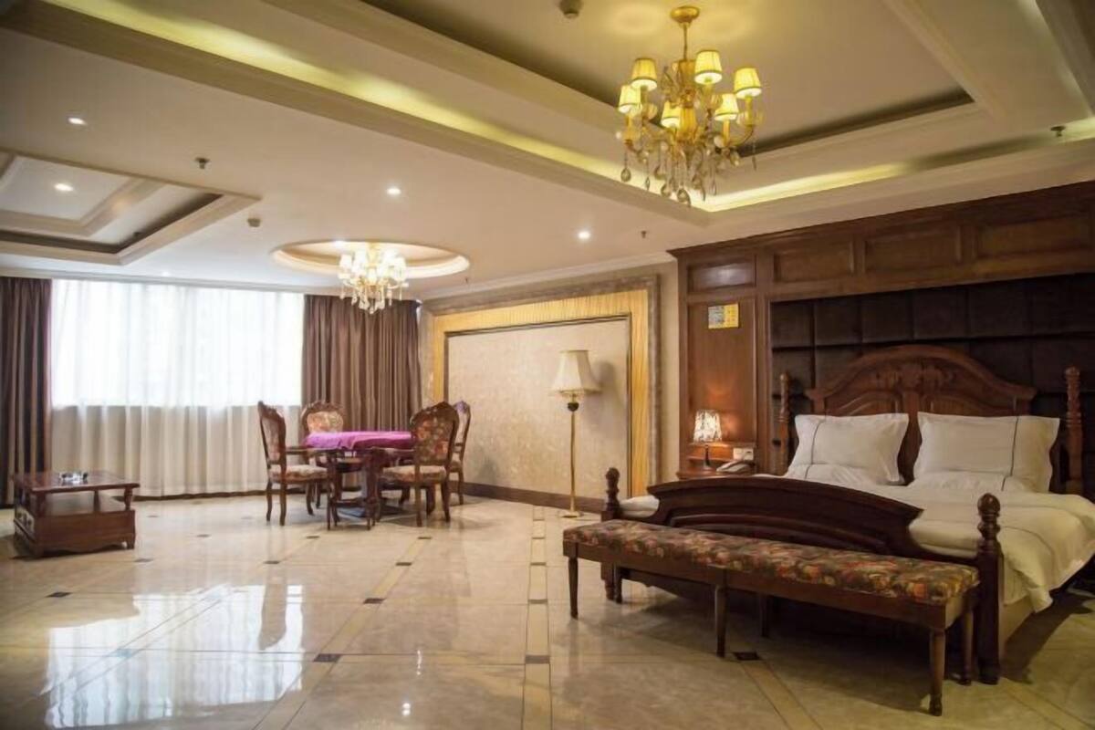 A hotel room with a bed, a bench, a desk with chairs, a chandelier, and a large window with curtains.