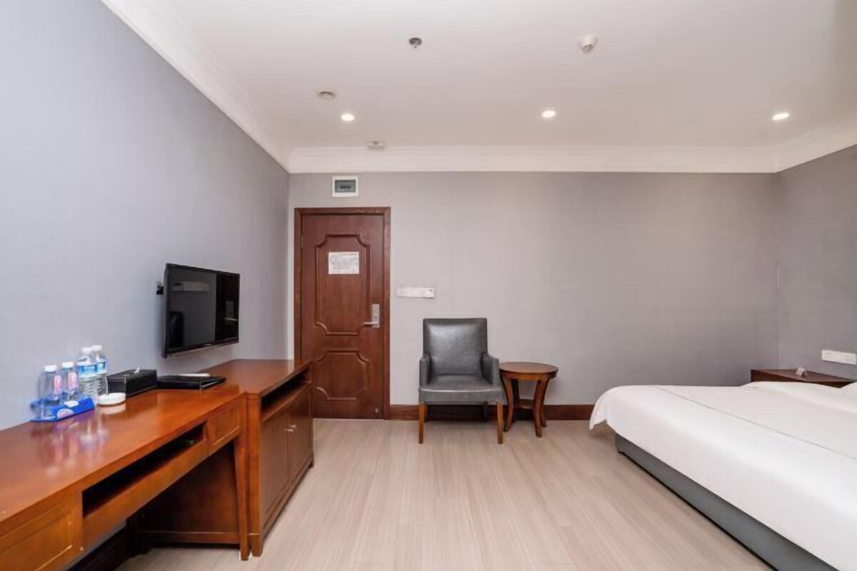 A hotel room with a bed, a desk with a TV, a chair, a small table, and a door.