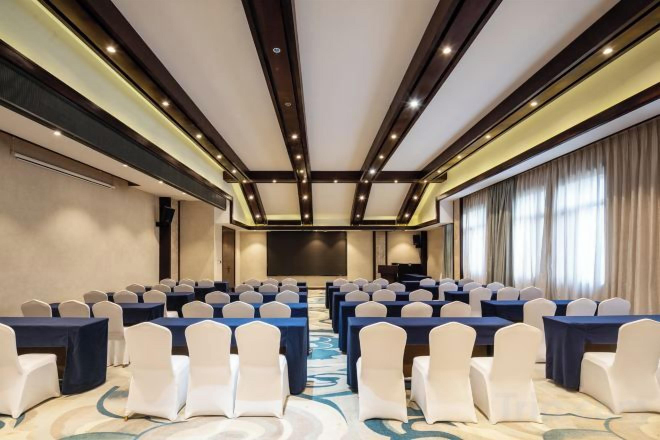 Un salón de conferencias con filas de mesas y sillas, una pantalla grande y suelo alfombrado.