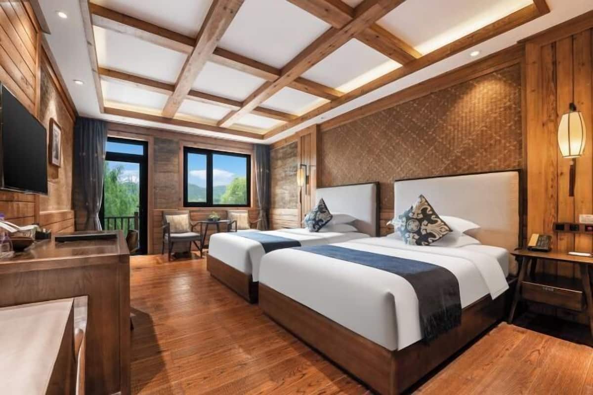 A hotel room with two beds, wooden flooring, a desk, and a large window offering a view of greenery.