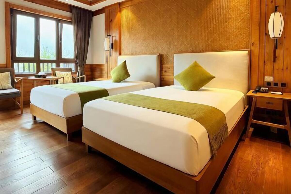 A hotel room with two beds, wooden flooring, a window with curtains, a desk, and a lamp.